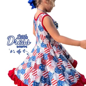May include: A red, white, and blue sleeveless dress with a firework print and a red ruffle trim. The dress is on a young girl with blonde hair and a blue bow.