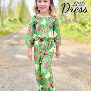 May include: A young girl wearing a green jumpsuit with a pattern of brown and tan shapes. The jumpsuit has long sleeves and pants, with a gathered waist. The text "Little Dress Shoppe" is visible in the upper right corner.