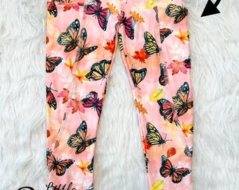 May include: Pink leggings with a butterfly and leaf print. The leggings have pockets on the sides. The text "Pockets" is written on the image.