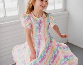 May include: A young girl wearing a rainbow and unicorn print dress with a white beaded necklace. The dress has a ruffled neckline and short sleeves. The girl is smiling and twirling in the dress.