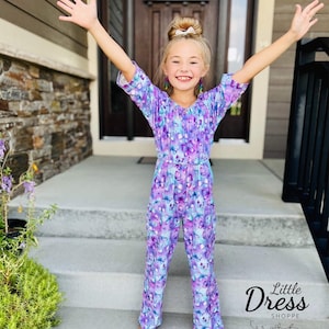 May include: A young girl wearing a purple and blue jumpsuit with a butterfly print. The jumpsuit has a ruffled neckline and 3/4 sleeves. The girl is standing on a set of stairs with her arms raised. The text "Little Dress Shoppe" is visible in the bottom right corner of the image.