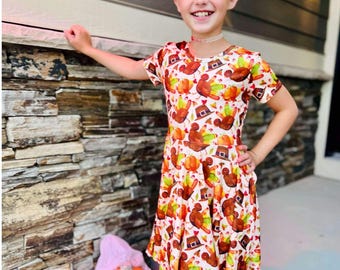 May include: A young person wearing a white dress with a Thanksgiving turkey print. The dress has a ruffled skirt and short sleeves. The person is standing in front of a brick wall and holding a small pumpkin.