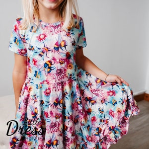 May include: A young girl wearing a short-sleeved dress with a watercolour print of bees and flowers. The dress has a ruffled hem and is a light blue, pink, and yellow colour.
