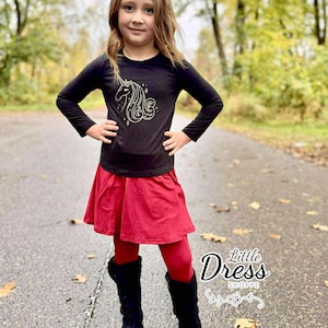 May include: A young girl wearing a black long-sleeved shirt with a white unicorn design, a red skirt, red leggings, and black boots. The image is taken outdoors on a wet road with trees in the background. The text "Little Dress Shoppe" is visible.