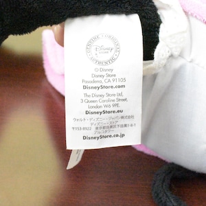 May include: A white tag with the Disney Store logo and text. The tag includes the Disney Store address in Pasadena, CA, and website information. The tag is attached to a pink and black plush toy.