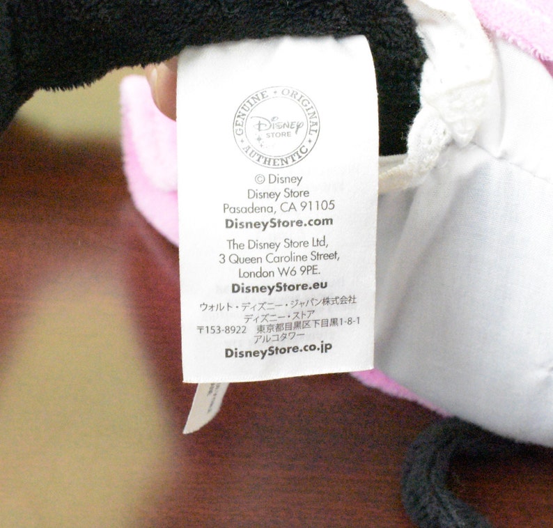 May include: A white tag with the Disney Store logo and text. The tag includes the Disney Store address in Pasadena, CA, and website information. The tag is attached to a pink and black plush toy.