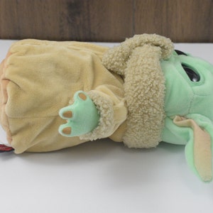 May include: A soft toy of a character from a popular science fiction franchise. The toy has a light green face, large black eyes, and tan ears. It is wearing a tan coat with a fluffy collar and has light green hands.