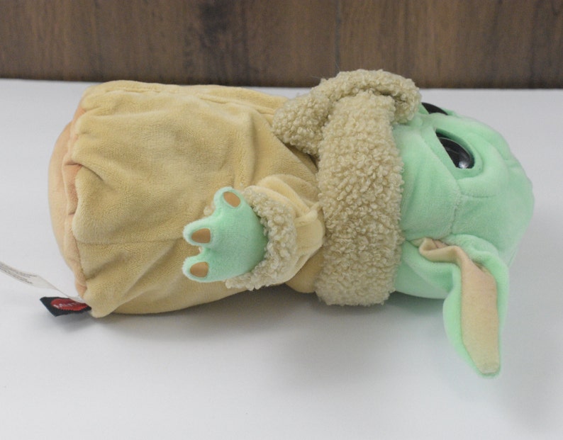 May include: A soft toy of a character from a popular science fiction franchise. The toy has a light green face, large black eyes, and tan ears. It is wearing a tan coat with a fluffy collar and has light green hands.