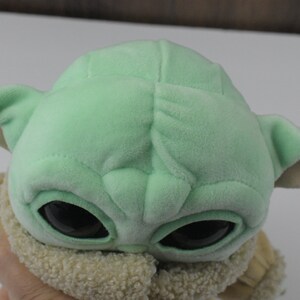 May include: A plush doll of a character with large black eyes, green skin, and large ears. The doll is wearing a tan and cream-coloured outfit. The doll is a popular character from a science fiction series.