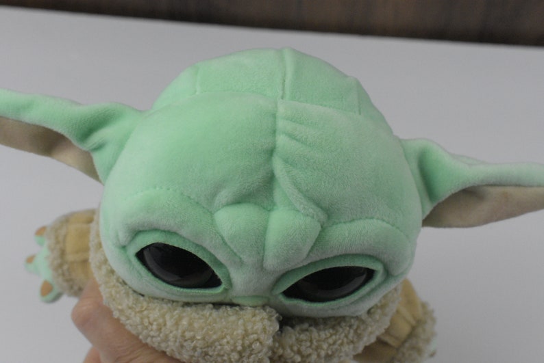 May include: A plush doll of a character with large black eyes, green skin, and large ears. The doll is wearing a tan and cream-coloured outfit. The doll is a popular character from a science fiction series.