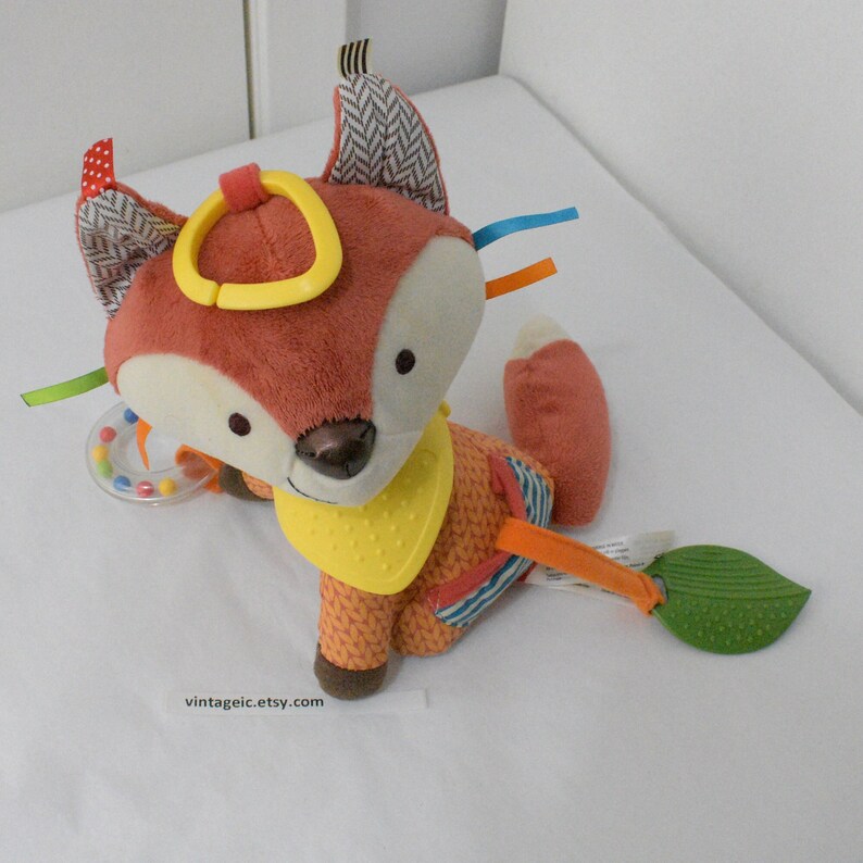 May include: A plush fox toy with a rust-colored body, white face, and patterned ears. It features a yellow teething ring, a yellow bib, and colorful ribbons. A green leaf-shaped teether is attached to the fox's tail. The toy is designed for babies.