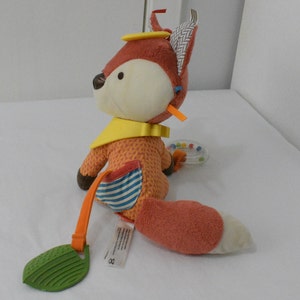 May include: A plush fox toy in shades of orange, cream, and brown, with a yellow bib and a striped pocket. The fox has a rattle and a green leaf-shaped teether attached. The toy is designed for babies and young children.