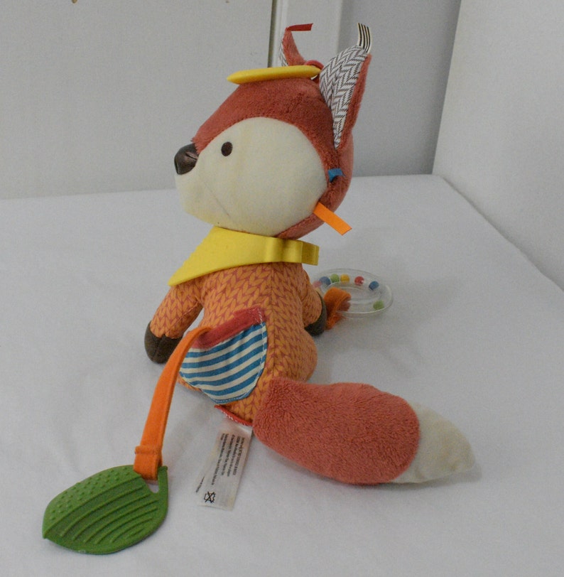 May include: A plush fox toy in shades of orange, cream, and brown, with a yellow bib and a striped pocket. The fox has a rattle and a green leaf-shaped teether attached. The toy is designed for babies and young children.