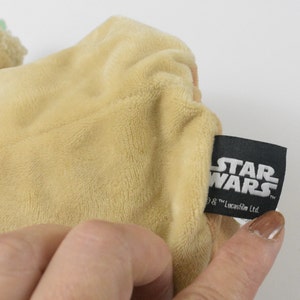 May include: A tan-coloured Star Wars character plush toy with a green head. The toy is made of soft, textured fabric. A black tag with the Star Wars logo is visible.
