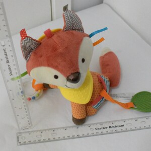 May include: A plush fox-shaped baby toy with a rattle and textured leaf. The fox has orange and cream fur, patterned ears, and a yellow bib. Colorful ribbons and a ring with small beads are attached. The toy is designed for infants.