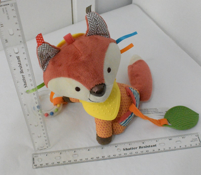 May include: A plush fox-shaped baby toy with a rattle and textured leaf. The fox has orange and cream fur, patterned ears, and a yellow bib. Colorful ribbons and a ring with small beads are attached. The toy is designed for infants.