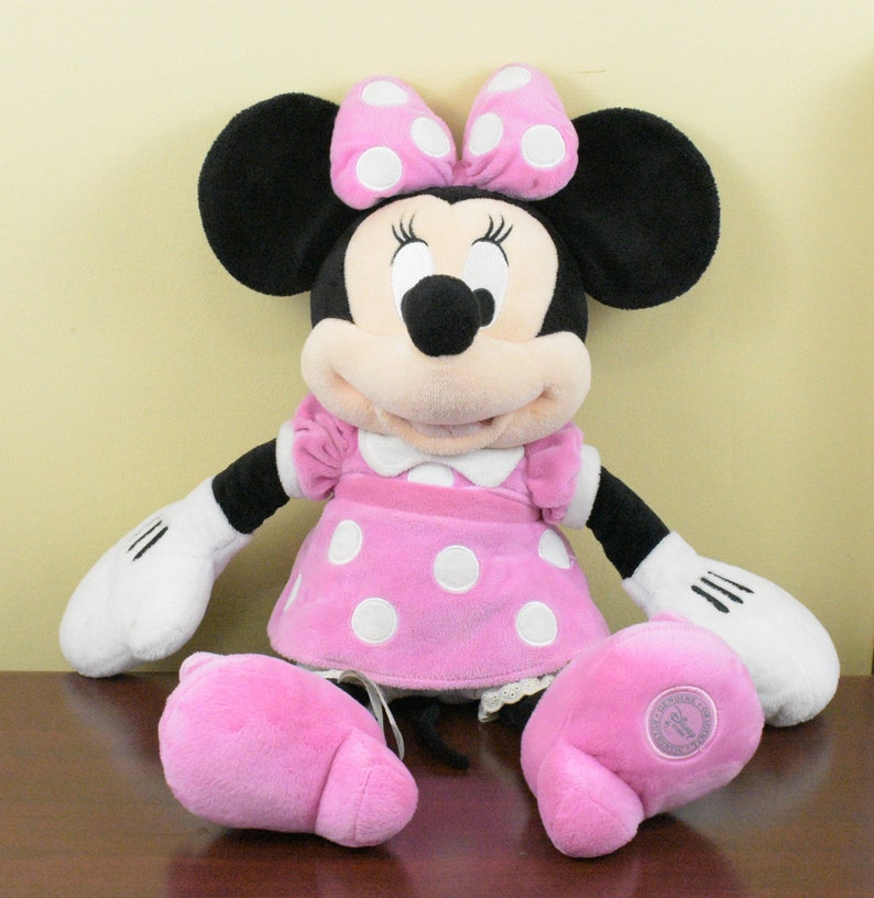 May include: A Minnie Mouse plush doll with a pink dress and bow, both adorned with white polka dots. The doll has black ears, a black nose, and white gloves. The doll's feet are also pink.