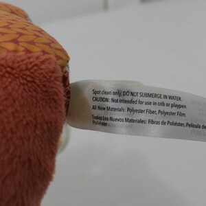 May include: Close-up of a plush toy with a peach-colored, textured exterior and a patterned orange and red top. A white tag is attached, with text indicating care instructions and materials: Polyester Fiber and Polyester Film.