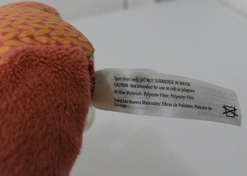 May include: Close-up of a plush toy with a peach-colored, textured exterior and a patterned orange and red top. A white tag is attached, with text indicating care instructions and materials: Polyester Fiber and Polyester Film.