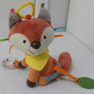 May include: A plush fox toy with a yellow bib, orange and brown fur, and a white face. The toy has a yellow loop, colorful ribbons, and a rattle. A green leaf-shaped teether is attached.