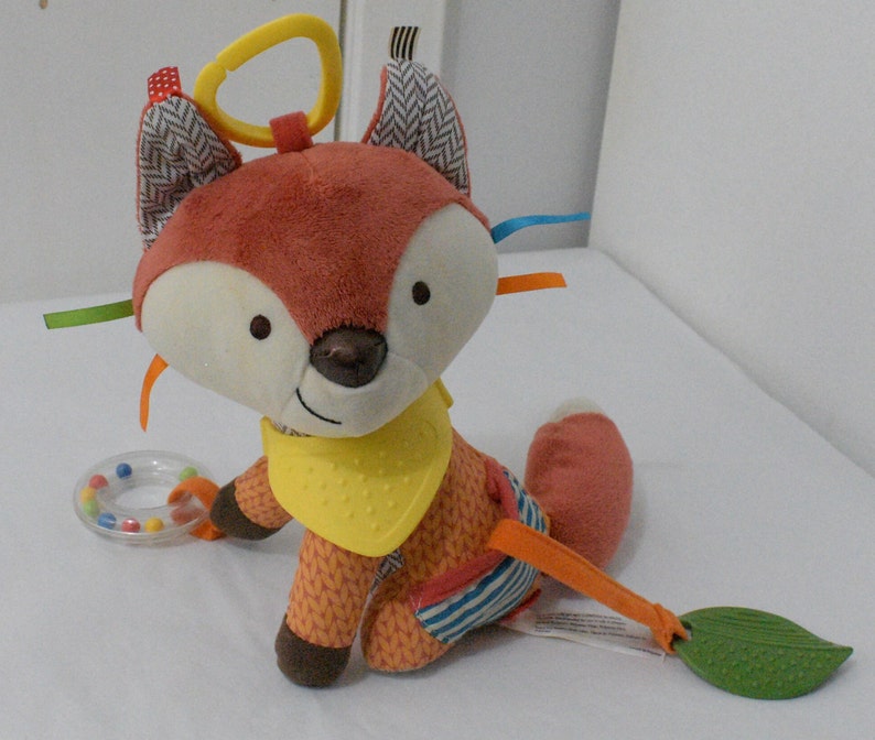 May include: A plush fox toy with a yellow bib, orange and brown fur, and a white face. The toy has a yellow loop, colorful ribbons, and a rattle. A green leaf-shaped teether is attached.
