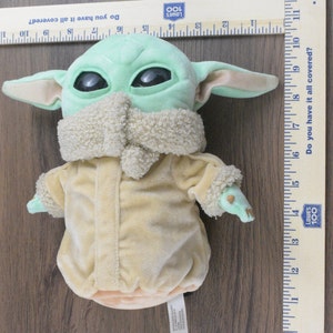 May include: A plush doll of a character with large black eyes, green skin, and large ears. The doll is wearing a tan coat with a fluffy collar and cuffs. A wooden ruler with cm measurements is next to the doll.
