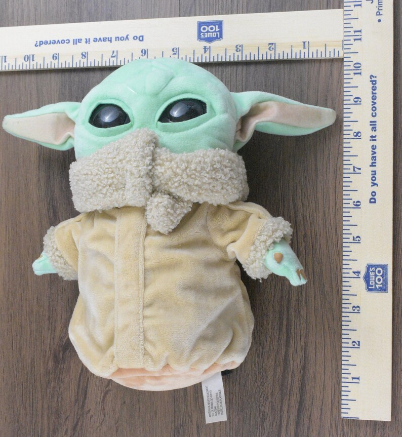 May include: A plush doll of a character with large black eyes, green skin, and large ears. The doll is wearing a tan coat with a fluffy collar and cuffs. A wooden ruler with cm measurements is next to the doll.