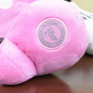 May include: Close-up of a pink plush toy foot with a circular "Genuine Disney Store Authentic Original" patch. The foot is part of a larger stuffed animal, with a white plush element visible in the background.
