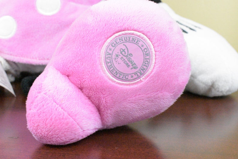 May include: Close-up of a pink plush toy foot with a circular "Genuine Disney Store Authentic Original" patch. The foot is part of a larger stuffed animal, with a white plush element visible in the background.