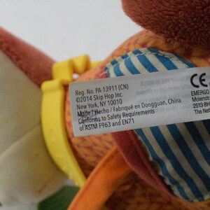 May include: A colorful plush toy with a tag featuring text including the brand name and safety information. The toy has orange, yellow, and blue accents. The tag includes the text "Reg. No. PA-13911 (CN)" and "&copy;2014 Skip Hop Inc."