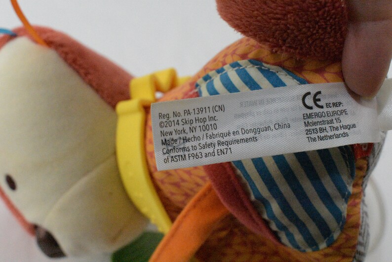 May include: A colorful plush toy with a tag featuring text including the brand name and safety information. The toy has orange, yellow, and blue accents. The tag includes the text "Reg. No. PA-13911 (CN)" and "&copy;2014 Skip Hop Inc."