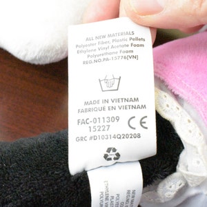 May include: A close-up of a tag with text, including "ALL NEW MATERIALS" and "MADE IN VIETNAM". The tag is attached to a plush toy with black, white, and pink fabric. The tag also includes a CE mark.