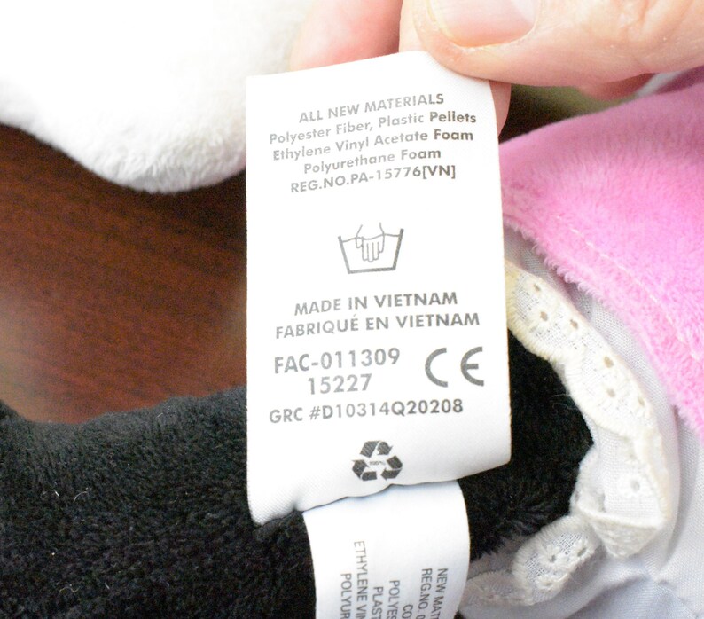 May include: A close-up of a tag with text, including "ALL NEW MATERIALS" and "MADE IN VIETNAM". The tag is attached to a plush toy with black, white, and pink fabric. The tag also includes a CE mark.