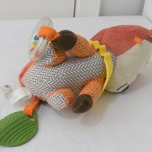 May include: A plush fox-shaped baby toy with a rattle and leaf-shaped teether. The fox has orange, brown, and cream-colored sections with a herringbone pattern on its body. The rattle contains colorful beads, and the teether is green.
