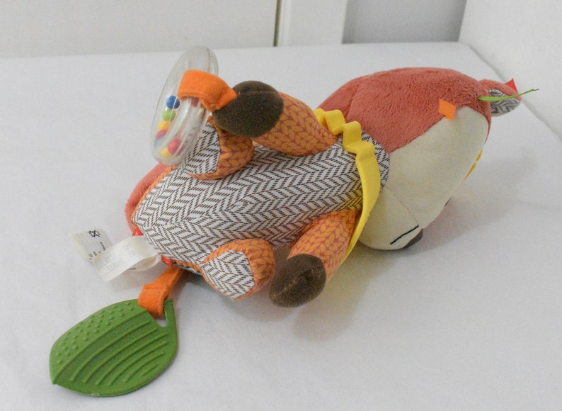 May include: A plush fox-shaped baby toy with a rattle and leaf-shaped teether. The fox has orange, brown, and cream-colored sections with a herringbone pattern on its body. The rattle contains colorful beads, and the teether is green.