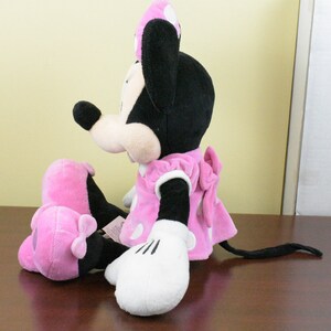 May include: A plush Minnie Mouse doll, featuring a black head and ears, a pink dress with white polka dots, and white gloves. The doll is sitting with its legs extended, showcasing a pink bow on the back of the dress.