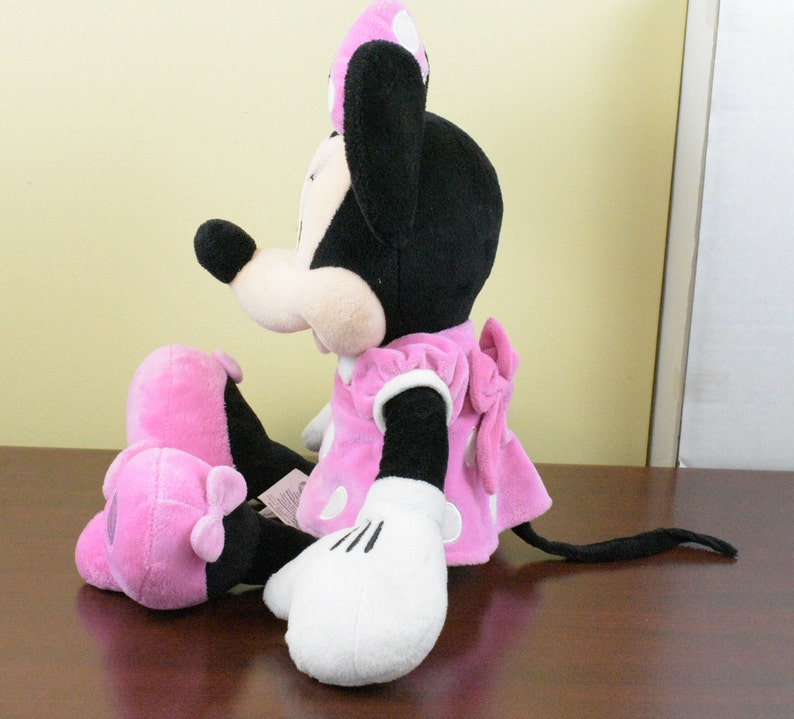 May include: A plush Minnie Mouse doll, featuring a black head and ears, a pink dress with white polka dots, and white gloves. The doll is sitting with its legs extended, showcasing a pink bow on the back of the dress.