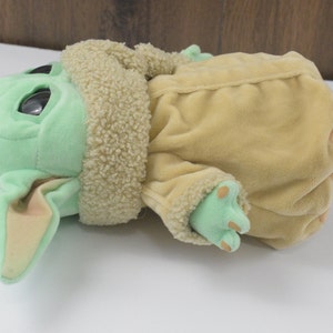 May include: A soft toy of a green-skinned character with large eyes and pointed ears. It is wearing a tan coat with a fluffy collar and has green feet. The toy is lying on its side.