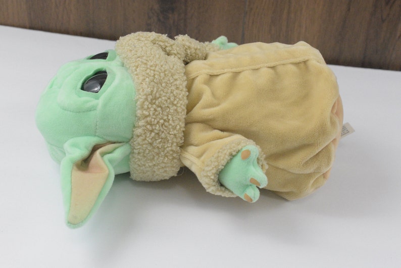 May include: A soft toy of a green-skinned character with large eyes and pointed ears. It is wearing a tan coat with a fluffy collar and has green feet. The toy is lying on its side.