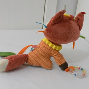 May include: A plush fox toy in shades of orange, brown, and cream, with a yellow ruffled collar and colorful ribbons. The fox has a rattle ring attached. The toy is designed for babies and young children.