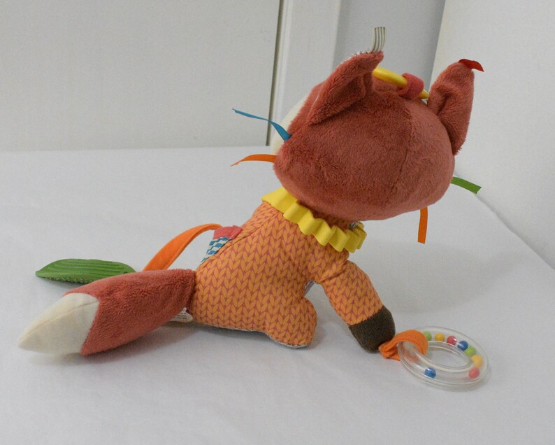 May include: A plush fox toy in shades of orange, brown, and cream, with a yellow ruffled collar and colorful ribbons. The fox has a rattle ring attached. The toy is designed for babies and young children.