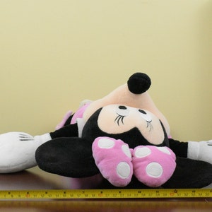 May include: A plush Minnie Mouse doll, lying on a brown surface. The doll features a black body, pink polka-dot shoes, and white gloves. The doll's head is pink with black ears and a black bow. A yellow measuring tape is in the foreground.