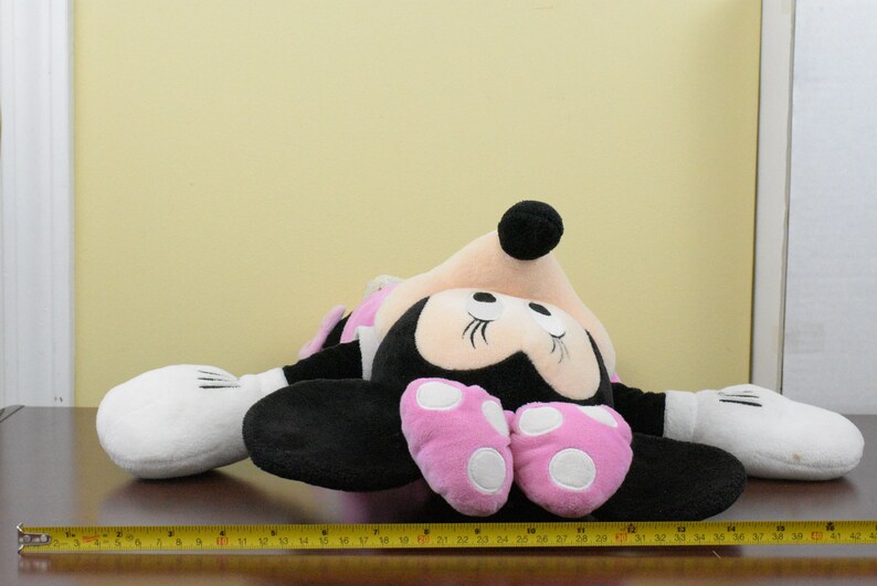 May include: A plush Minnie Mouse doll, lying on a brown surface. The doll features a black body, pink polka-dot shoes, and white gloves. The doll's head is pink with black ears and a black bow. A yellow measuring tape is in the foreground.
