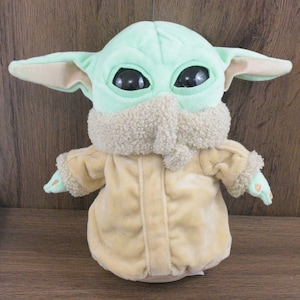 May include: A soft toy of a character with large black eyes, large ears, and a light green head. The character wears a beige coat with a fluffy collar and cuffs. The toy is on a brown wooden surface.