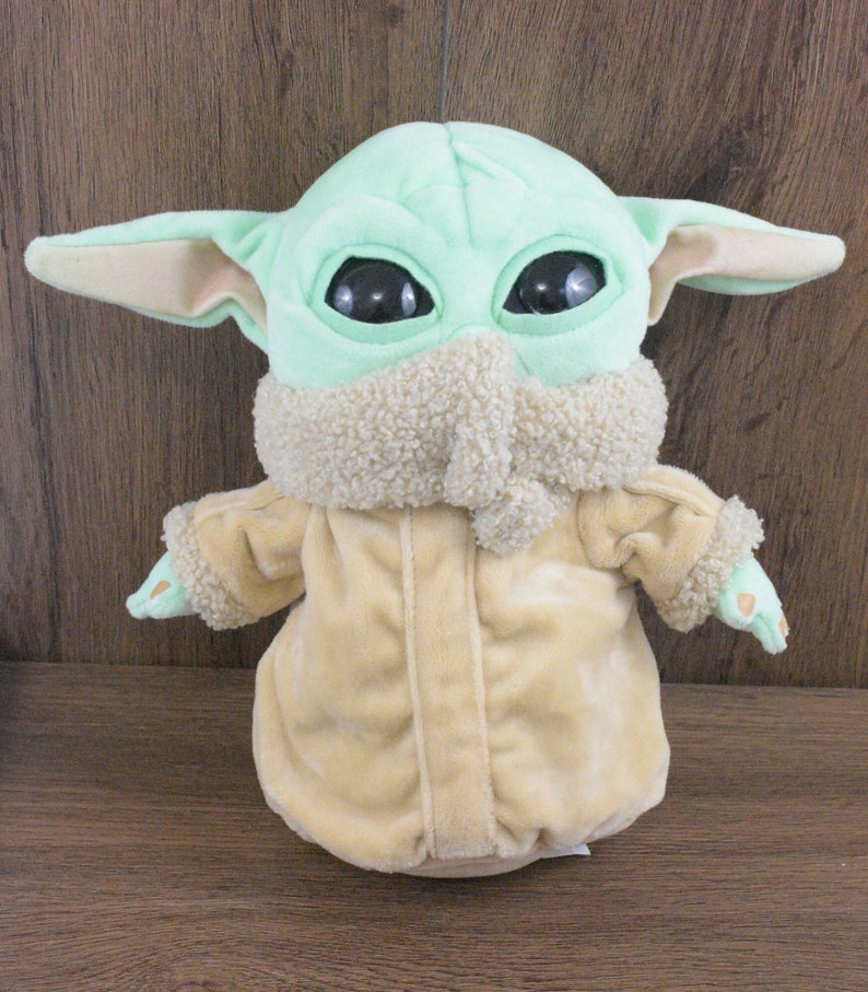 May include: A soft toy of a character with large black eyes, large ears, and a light green head. The character wears a beige coat with a fluffy collar and cuffs. The toy is on a brown wooden surface.