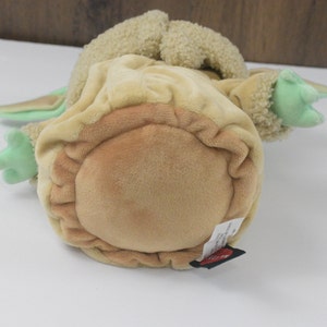 May include: A soft toy of a character from a popular science fiction series. The toy is mainly tan and light green, with large ears and small hands. The toy is lying on its back.