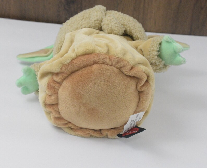 May include: A soft toy of a character from a popular science fiction series. The toy is mainly tan and light green, with large ears and small hands. The toy is lying on its back.