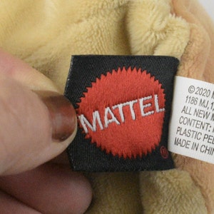 May include: A close-up of a toy tag featuring the MATTEL logo in a red circle on a black label. The tag also includes text about the toy's content and origin. The plush toy is a light tan colour.