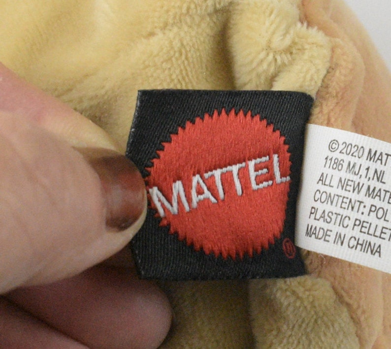 May include: A close-up of a toy tag featuring the MATTEL logo in a red circle on a black label. The tag also includes text about the toy's content and origin. The plush toy is a light tan colour.
