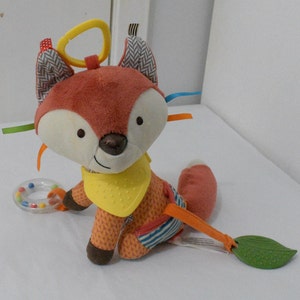 May include: A plush fox baby toy with a yellow bib, orange and white patterned body, and a leaf-shaped teether. The toy has a yellow ring for hanging and colorful ribbons. A small rattle is also included.